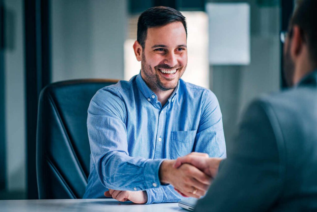 two smiling businessmen shaking hands while sitting office desk Easy Resize.com 1